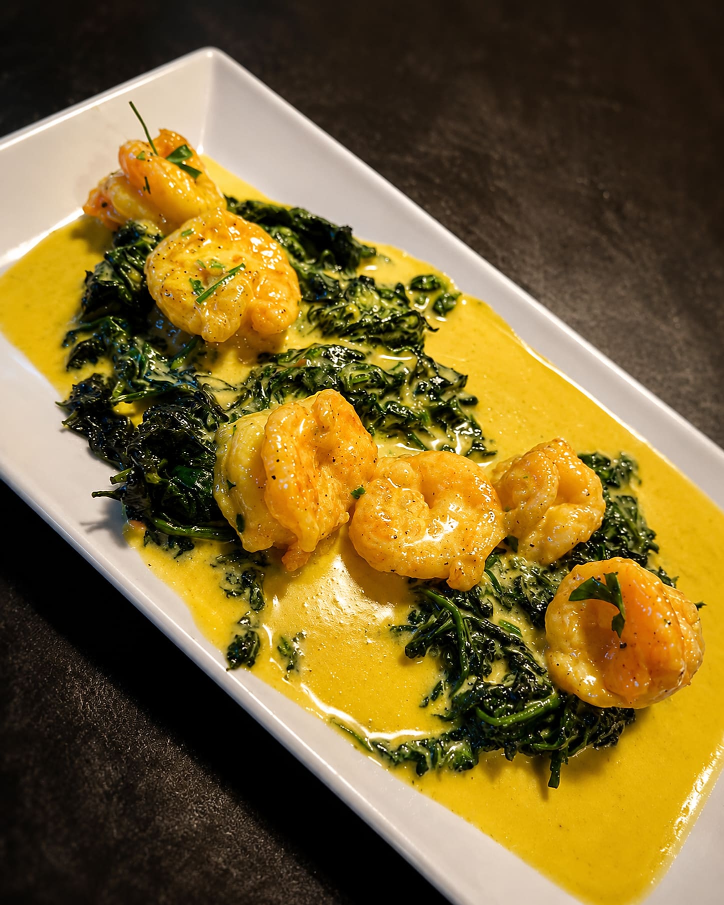 Shrimp in a saffron-coloured sauce laid along a bed of sautéed broccoli rabe on a long rectangular plate.