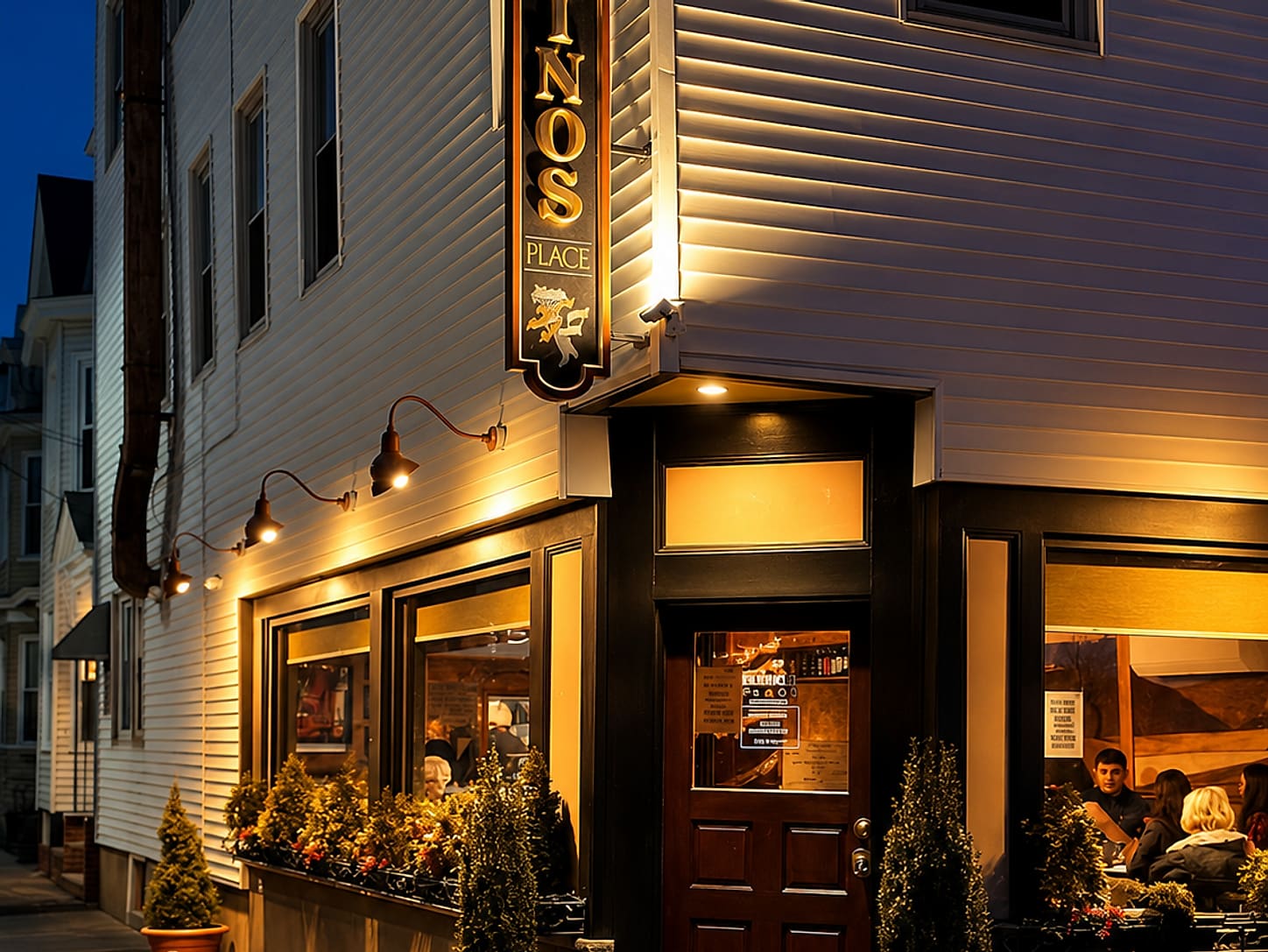Rino’s Place at night on Saratoga Street — a hanging sign lettered RINO’S PLACE over a gold lion, wall lamps along white clapboard, warm light in the windows and people at the tables inside.